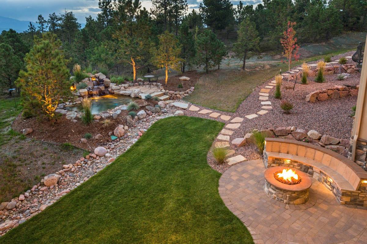 Twilight backyard with a lit fire pit, paver patio, retaining-wall bench, waterfall, and uplit pines — JR Landscaping homepage photo
